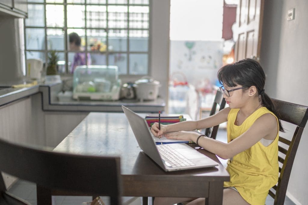 young girl attending online class at the kitchen dining table while her grandmother preparing food in kitchen