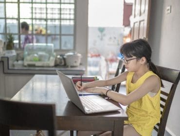 young girl attending online class at the kitchen dining table while her grandmother preparing food in kitchen