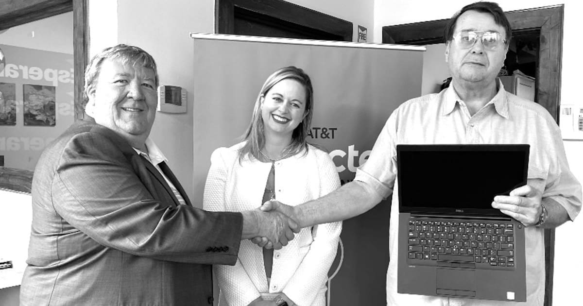 Two people sharing hands, with a woman standing behind them. The person on the right is holding a laptop.