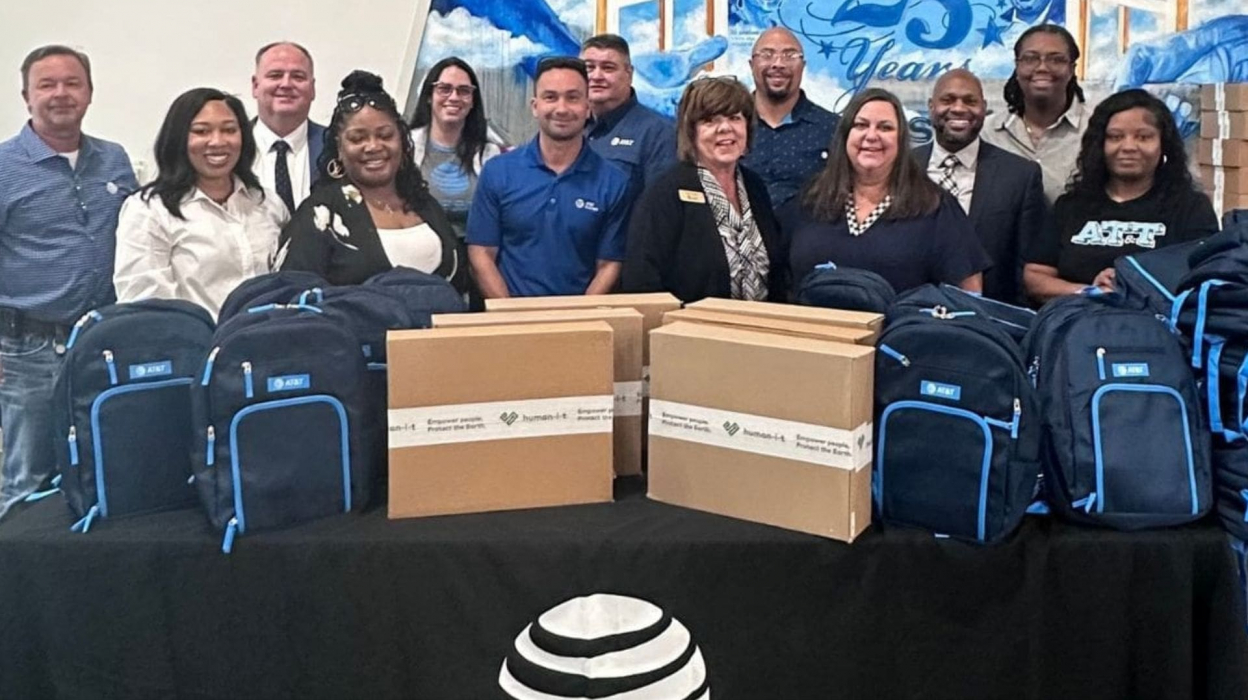 A group of 13 people standing behind a table which has AT&T branded backpacks on it.