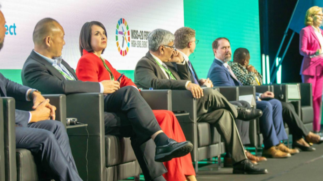 An image of 7 people sitting on stage, as part of a panel, with the emcee at the podium
