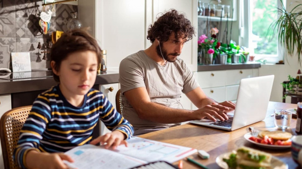 An adult and child at a kitchen table. The adult is working on a laptop with headphones in. The child is flipping through a book.