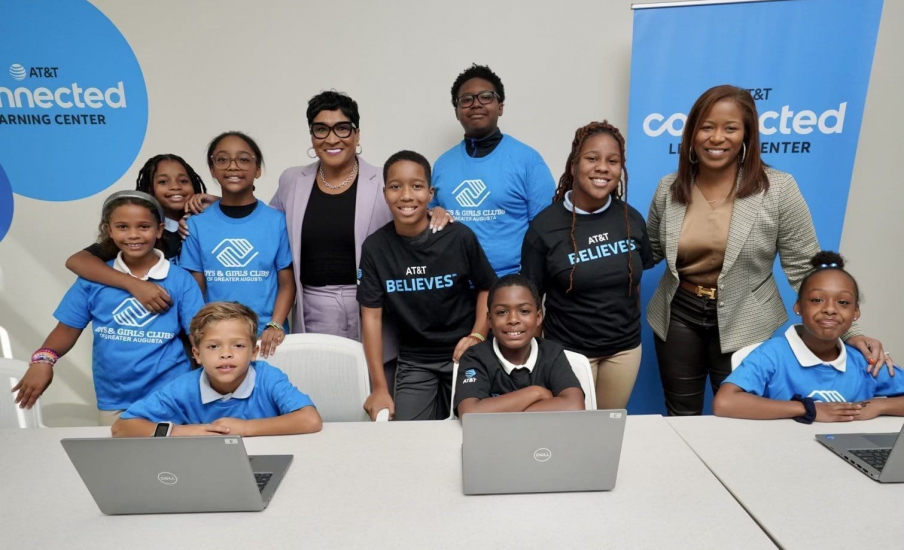 A group of kids and adults standing around a table smiling with two laptops in front of them.