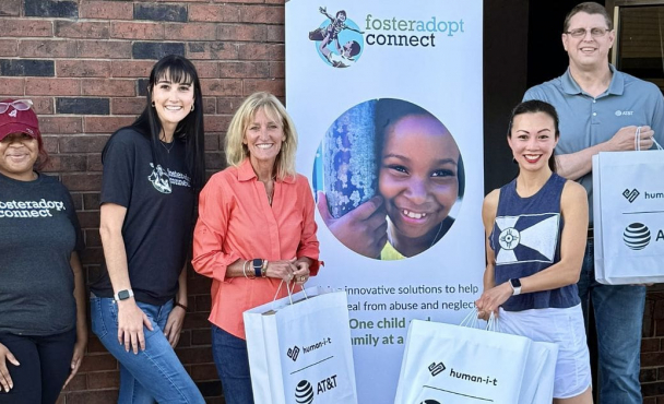 A group of 5 people outside a brick building with a foster adopt connect banner in background holding white bags with the human-it and AT&T logos on it