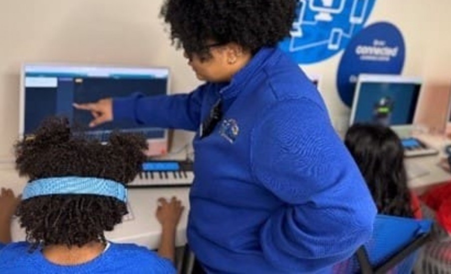 A adult woman talking to a student with headphones on at a computer.