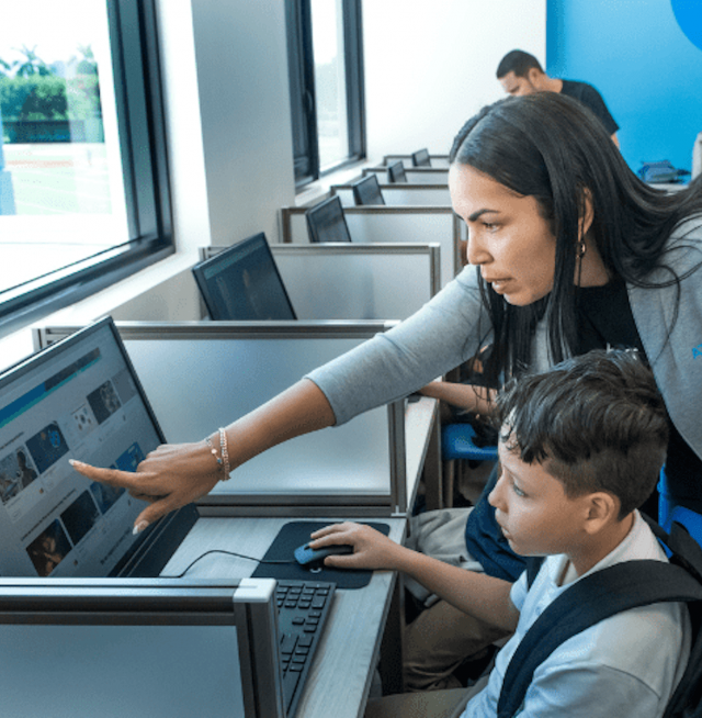 A boy sitting at a computer with a woman pointing at a computer screen
