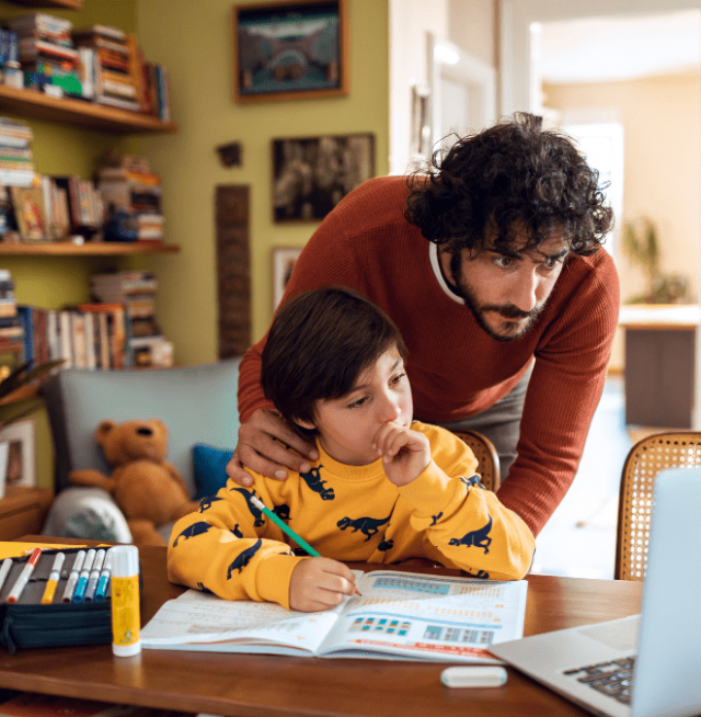 An adult with a young child sitting in front of a laptop at a table, with the child using a pencil to fill in a notebook.