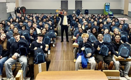 A man standing in the middle of a room waving at the camera, children facing the camera in matching sweatshirts with matching backpacks